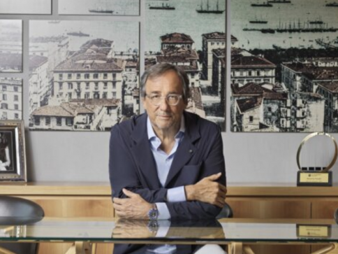 A man with glasses and a blue blazer sits at a wooden desk, arms folded. Behind him are framed historic cityscape photographs and a trophy.