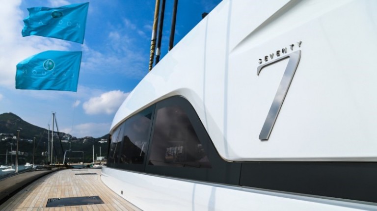 Close-up of a luxury yacht moored at a marina, with the number 7 and the word Seventy on its side. Two blue flags are flying above, and hills are visible in the background under a partly cloudy sky.