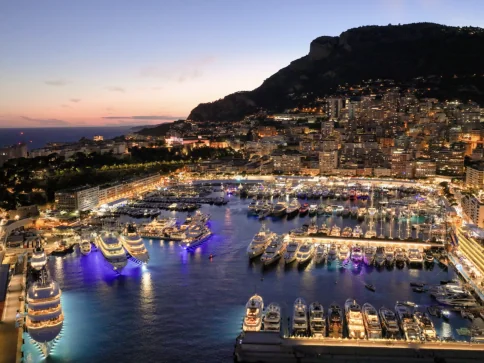 Aerial view of a marina at dusk with many yachts moored, city buildings lit up along the shoreline, and a mountain in the background under a twilight sky.