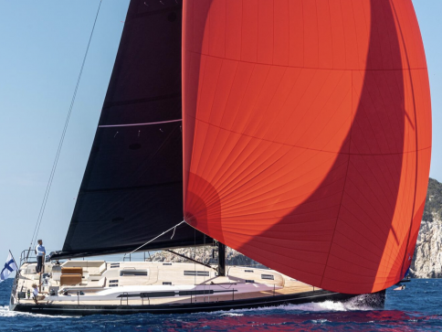 A modern sailboat with a large red spinnaker sail glides on blue ocean water near rocky cliffs, with a person standing at the helm and a flag flying at the stern.