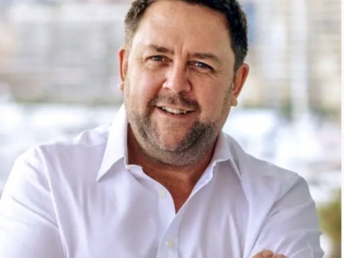A man with short dark hair and a beard, wearing a white collared shirt, stands smiling with arms folded. The background is blurred, showing outdoor scenery with buildings.