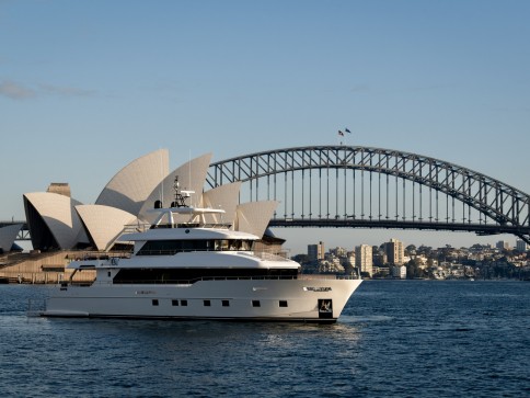A white yacht sails on Sydney Harbour with the Sydney Opera House and Harbour Bridge in the background under a clear blue sky.
