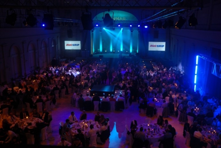 Large banqueting hall filled with people seated at round tables, dimly lit with blue stage lights. A stage with spotlights and two projection screens displaying the AWLGRIP logo are visible at the front.