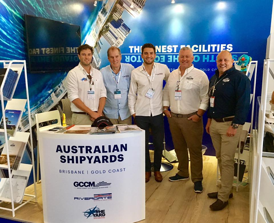 Five men stand smiling at an exhibition stand for Australian Shipyards, featuring promotional materials and a large backdrop with images of water and ships. The stand highlights locations in Brisbane and the Gold Coast.