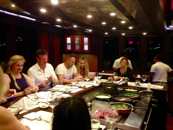 A group of people sit around a teppanyaki grill table in a restaurant, watching a chef cook. Plates, glasses, and food ingredients are arranged on the grill and table. The atmosphere is lively and sociable.