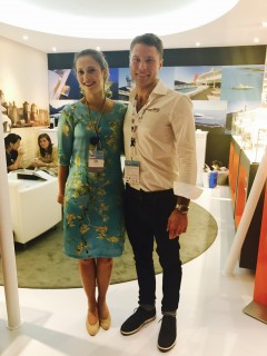 A woman in a blue floral dress and a man in a white shirt and dark trousers stand together smiling at an indoor event, with people seated in the background and aviation images on the walls.