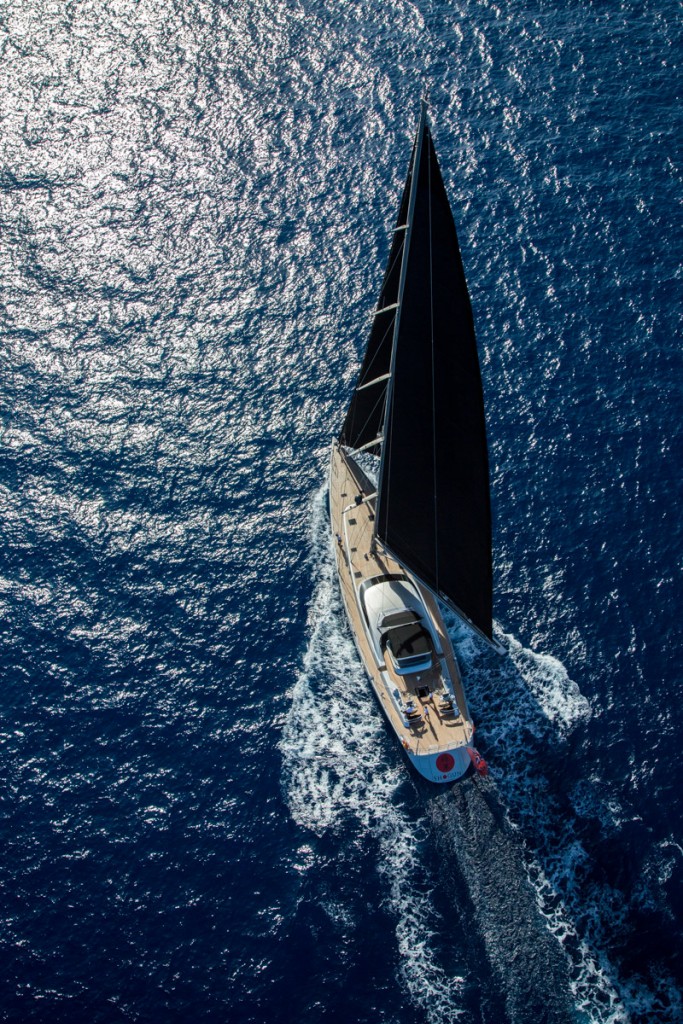 Aerial view of a sleek yacht with a dark sail cutting through deep blue ocean water, leaving a white wake behind as sunlight shimmers on the sea’s surface.