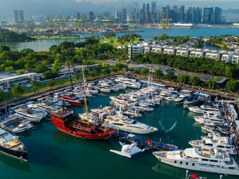 Aerial view of a marina filled with numerous yachts and boats, with a large red ship standing out among them. City skyline, green spaces, and water are visible in the background under a partly cloudy sky.