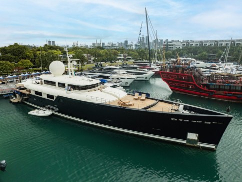 A large, modern black yacht moored at a marina alongside several other boats, with a city skyline and trees visible in the background under a partly cloudy sky.