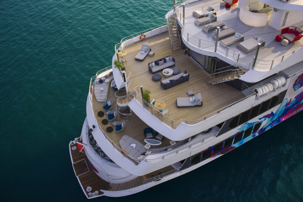 Aerial view of a luxury yacht with multiple decks featuring outdoor lounge areas, sunbeds, and modern seating, floating on calm turquoise water.