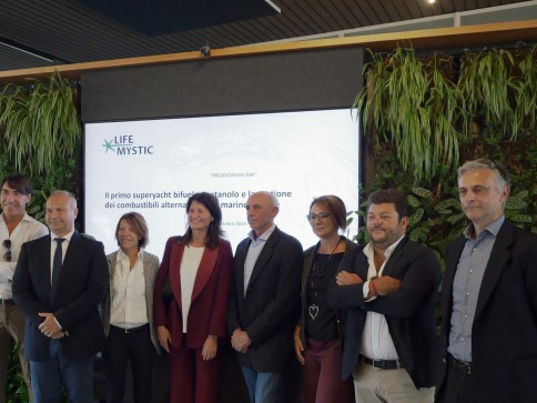 Eight people stand in front of a screen displaying the Life Mystic logo and text at a formal event, with green plants covering the wall behind them. They are dressed in business attire and smiling.