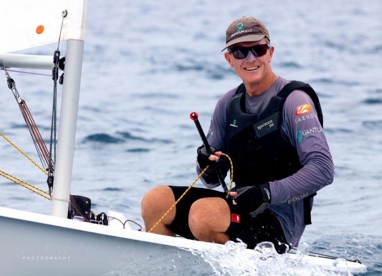 A smiling man wearing sunglasses, a cap, and sailing gear sits in a small sailing boat on the water, holding a rope and tiller, with light waves splashing against the boat.