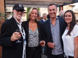 Four adults stand together at an outdoor social event, smiling at the camera. Three are holding drinks. They are dressed in business-casual attire with buildings visible in the background.