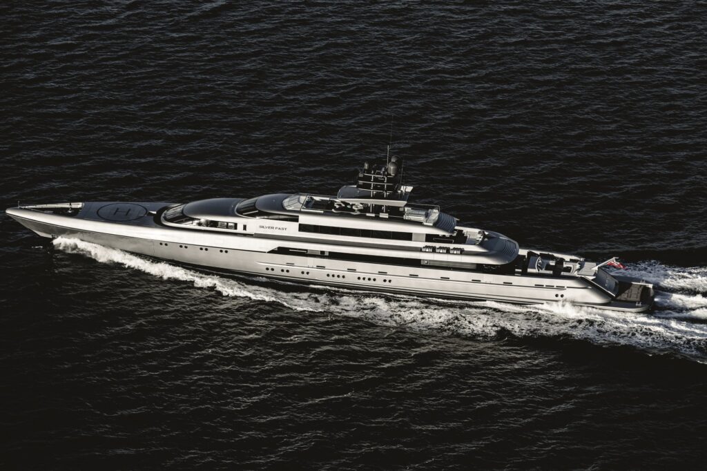 A sleek, modern luxury yacht cruises through dark blue ocean waters, leaving a white wake behind. The yacht has a helipad on the bow and multiple decks, with a flag at the stern.