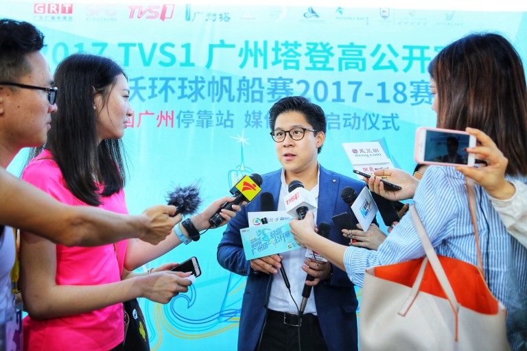 A man in a suit and glasses speaks to multiple reporters holding microphones and recording devices, with a blue event backdrop behind him containing Chinese text and logos.