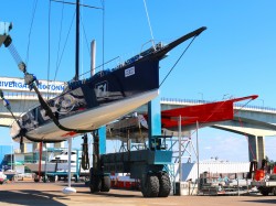 A large sailboat is suspended by a blue boat hoist at a marina, with another red boat in the background and a bridge overhead on a sunny day.