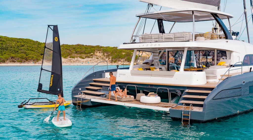 A family enjoys a sunny day on a luxury catamaran yacht anchored near a beach, with adults relaxing on the deck and children playing on paddleboards and a small sailing boat in the clear blue water.