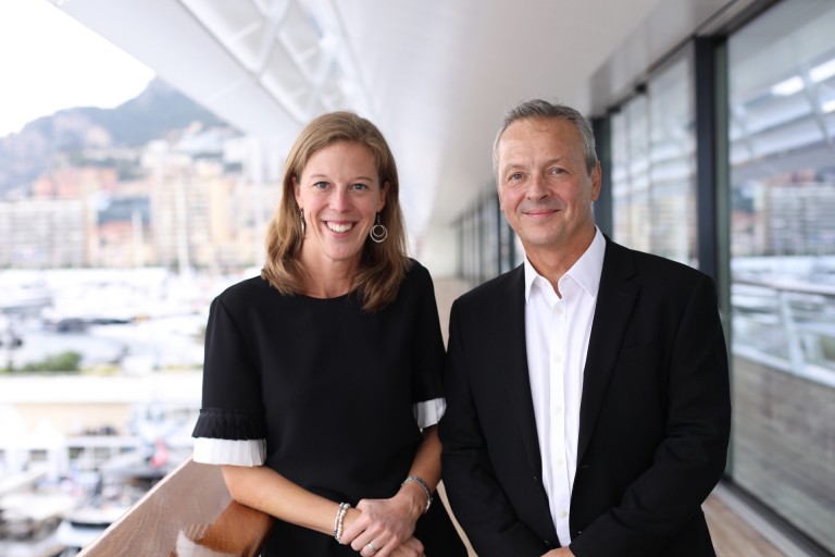 A woman and a man, both in business attire, stand side by side and smile at the camera on a balcony with a cityscape and marina in the background.