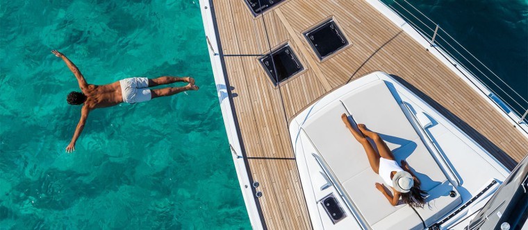 A person swims in clear turquoise water beside a yacht, whilst another person in a white swimming costume and hat relaxes on the yacht’s deck, enjoying the sun.