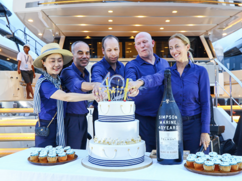 Five people in matching blue outfits stand on a yacht, smiling as they cut a large white cake together. Cupcakes surround the cake, and a giant wine bottle labelled Simpson Marine 40 Years stands beside them.