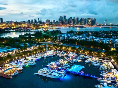 A marina filled with yachts and boats illuminated by colourful lights, with a city skyline and high-rise buildings in the background under a cloudy evening sky. Trees and waterfront homes line the area.
