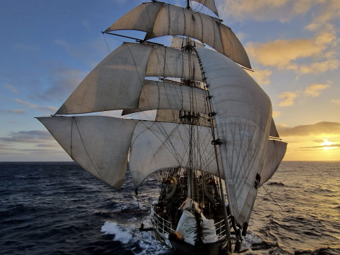 A tall sailing ship with large white sails moves across the open sea at sunset, with golden light illuminating the sky and clouds in the background.