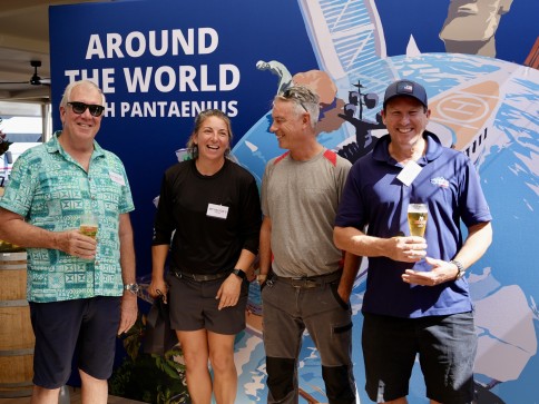 Four smiling people stand together holding drinks in front of a blue “Around the World with Pantaenius” backdrop featuring nautical graphics. They appear relaxed and friendly, enjoying the event.