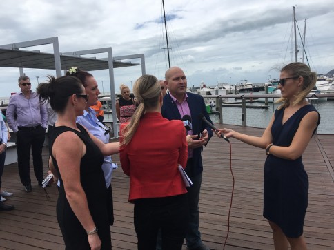 A man is being interviewed by three women holding microphones and a recorder on a wooden quay, with yachts and boats in the background. Several other people stand nearby under a cloudy sky.
