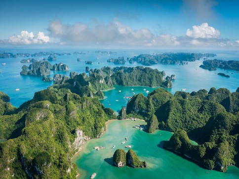 Aerial view of a turquoise bay dotted with lush, green limestone islands and rocky islets under a partly cloudy sky, with boat traffic visible in the water.