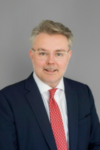 A man with grey hair and glasses, wearing a dark suit, white shirt, and red patterned tie, poses in front of a plain grey background.