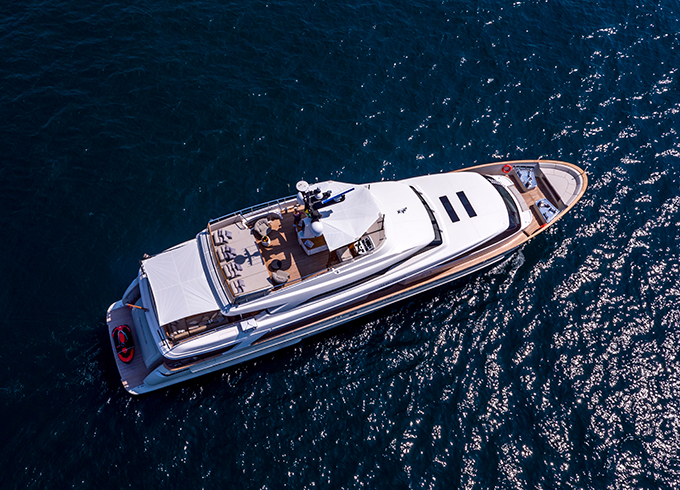 Aerial view of a luxurious white yacht cruising on deep blue water, with sun loungers and shaded seating areas visible on deck. The sunlight reflects off the calm sea around the vessel.