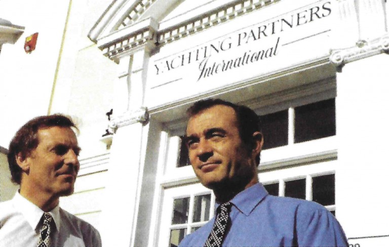 Two men in dress shirts and ties stand outside a white building with a sign above the entrance that reads “Yachting Partners International.” Both men are looking in different directions and smiling slightly.