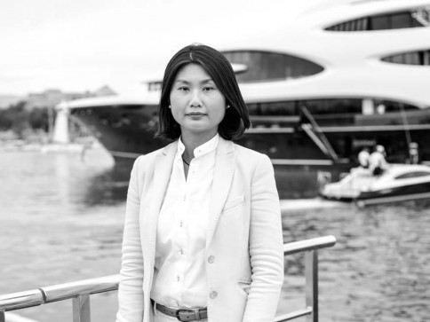 A woman in a light-coloured suit stands by a railing near a waterfront, with a large yacht and people on a smaller boat visible in the background. The image is in black and white.