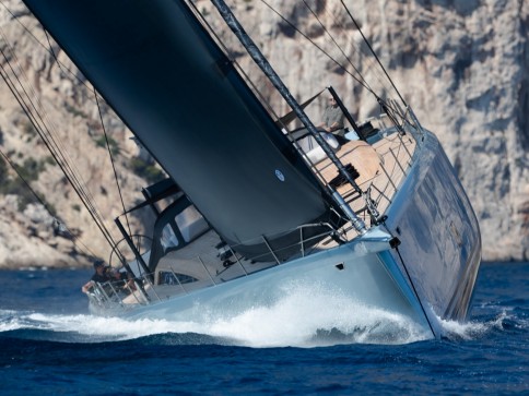 A sleek sailboat with a large dark sail cuts through blue ocean waves at an angle, creating white spray. Rocky cliffs are visible in the background under bright sunlight.