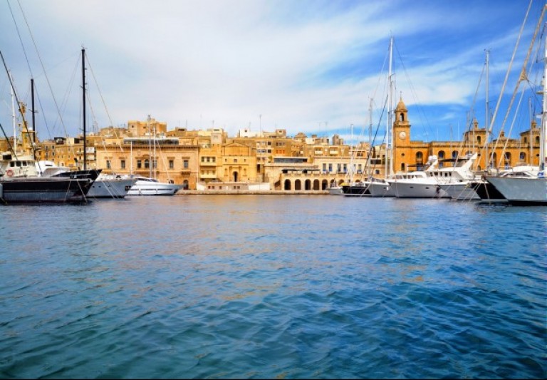 A marina with several yachts and sailboats moored in front of historic yellow stone buildings and a clock tower under a blue sky with wispy clouds. The water reflects the boats and waterfront architecture.