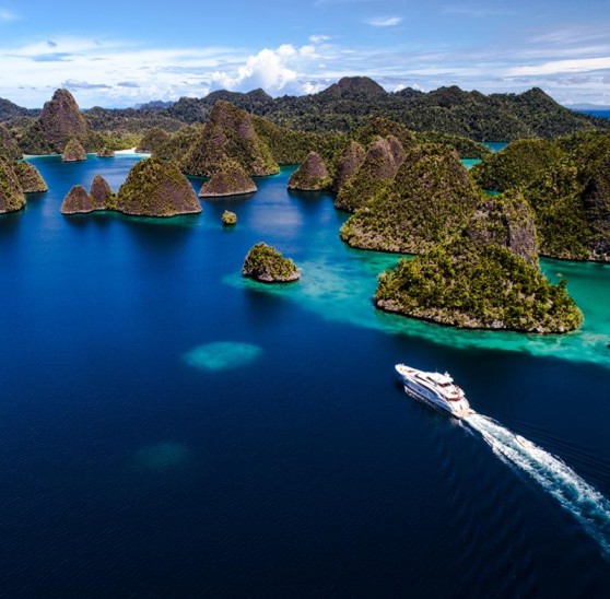 A white boat sails through turquoise water surrounded by lush, green, rocky islands and blue sky, creating a scenic tropical landscape.