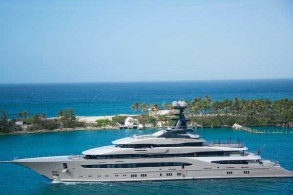 A large, modern luxury yacht sails on clear blue water near a tropical coastline with palm trees and a sandy beach under a bright, sunny sky.
