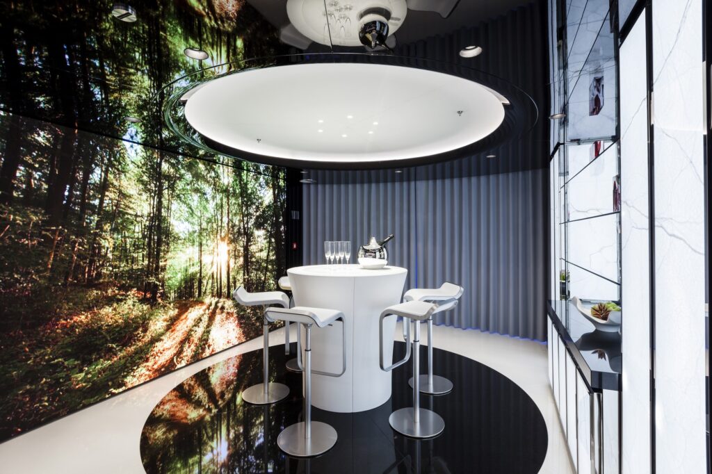 A modern, futuristic room with a circular white table surrounded by four white bar stools. A curved wall displays a forest image, and a curved partition adds contrast. Champagne and glasses sit on the table under a large circular ceiling light.