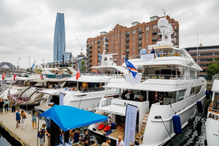 Luxury yachts moored at a marina during a boat show, with people walking along the quay. Modern buildings and a tall skyscraper are visible in the background under a cloudy sky.