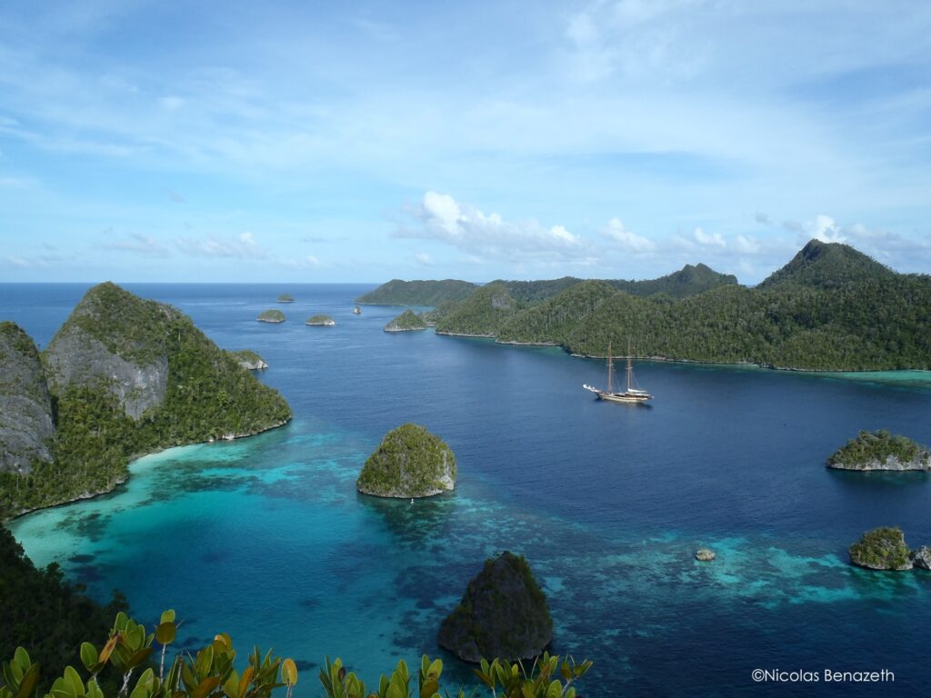 A scenic view of Raja Ampat islands in Indonesia, with lush green islets surrounded by clear blue water. A large sailing boat is anchored among the islands under a partly cloudy sky.
