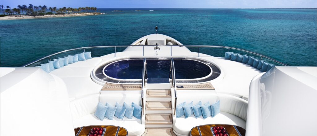 A luxury yacht deck with a circular pool, surrounded by white seating and blue cushions, overlooks clear turquoise water and a distant island with palm trees under a blue sky.