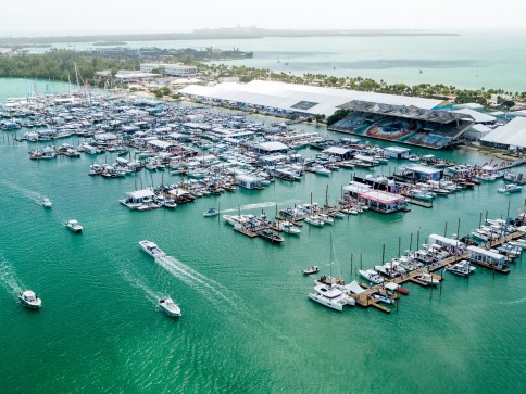 Aerial view of a marina with numerous boats and yachts moored along piers on turquoise water. Several small boats are cruising, and large white buildings and palm trees are visible in the background.