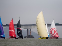 Several sailboats with colourful sails race on the water, with a large bridge and vehicles visible in the background under an overcast sky.