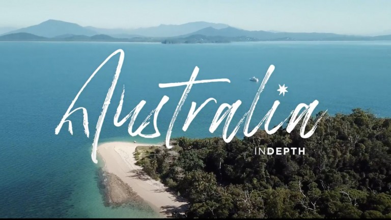 Aerial view of a lush, green coastline with clear blue water and distant mountains, overlaid with the words Australia INDEPTH in large, cursive white text.