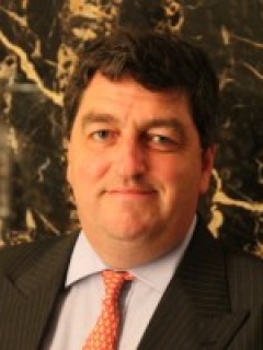 A man with dark hair wearing a dark pinstripe suit, light blue shirt, and orange patterned tie, standing in front of a dark marble background.