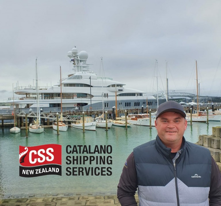 A man in a cap and waistcoat stands by a harbour with yachts and boats moored behind him. The sky is overcast. The Catalano Shipping Services New Zealand logo is displayed in the lower left corner of the image.