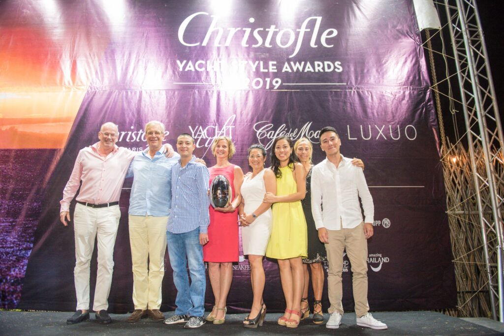 A group of eight people stand smiling in front of a banner for the 2019 Christofle Yacht Style Awards; one woman in the centre holds a trophy. The setting appears festive and is outdoors at night.