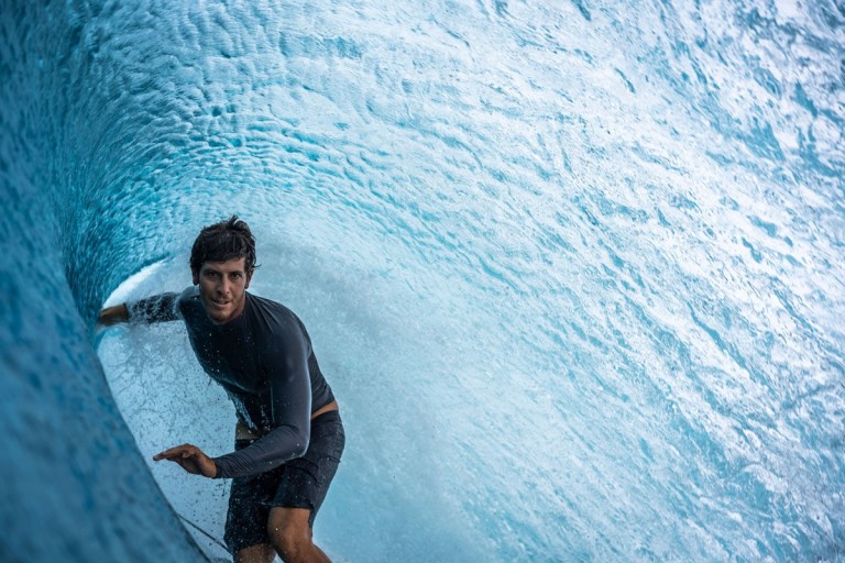 A surfer in a black wetsuit rides inside the barrel of a turquoise wave, with water curling overhead and sunlight filtering through.