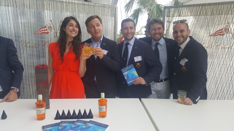 Five people, four men in suits and one woman in a red dress, stand smiling behind a table with bottles, drinks, and brochures. They appear to be at an outdoor event with palm trees in the background.