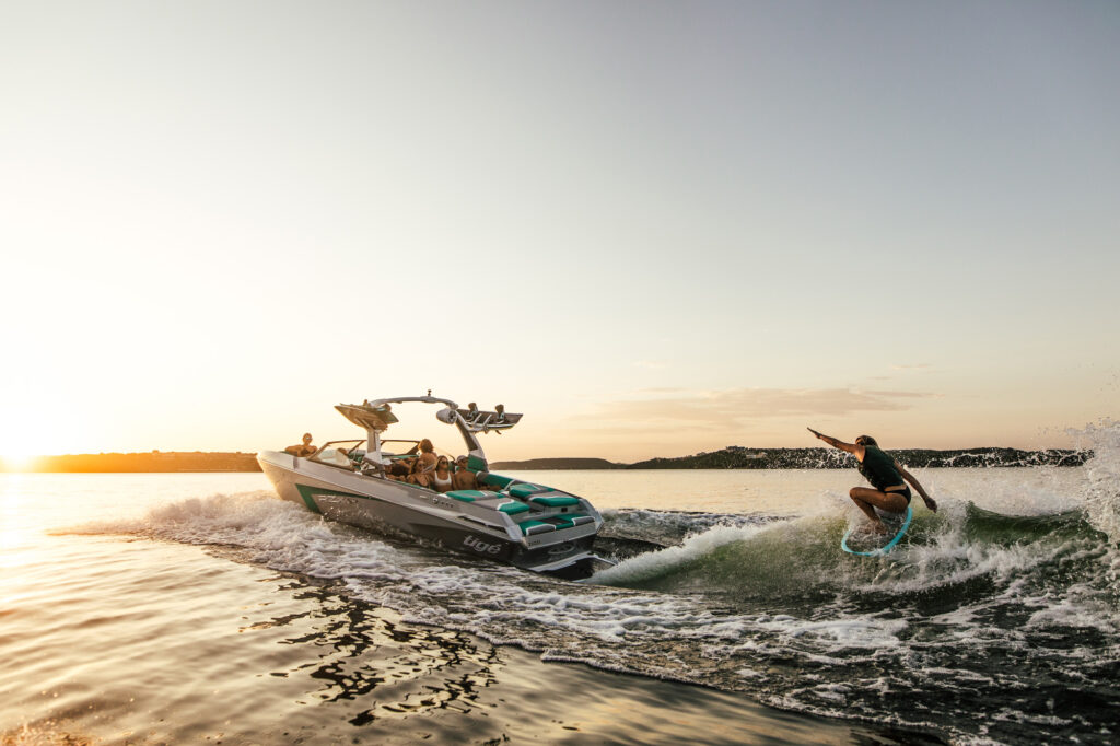 A person surfs on the wake behind a boat full of people during sunset on a calm lake, with hills visible in the background and the sky softly lit.